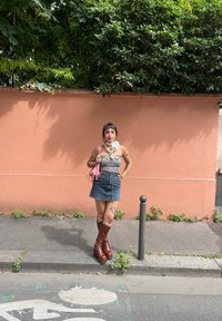 Δερμάτινη μίνι φούστα, γκρι τοπ χωρίς straps, ροζ τσάντα, χοντρά καστανά μποτάκια μέχρι το γόνατο, μεγάλο κολιέ, με φόντο ένα γκράφιτι και πράσινα στοιχεία.