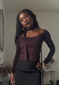 Top burdeos de manga larga con diseño de escote barco y detalle de lazo alrededor del cuello, combinado con una falda negra ajustada. Se observan accesorios dorados.