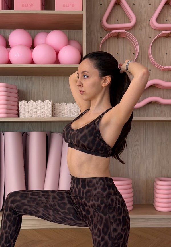 Nő tigrismintás edzőruhában hátrafogja a haját egy fitneszstúdióban, rózsaszín tornalabdákkal, szőnyegekkel és felszereléssel a fa polcokon.