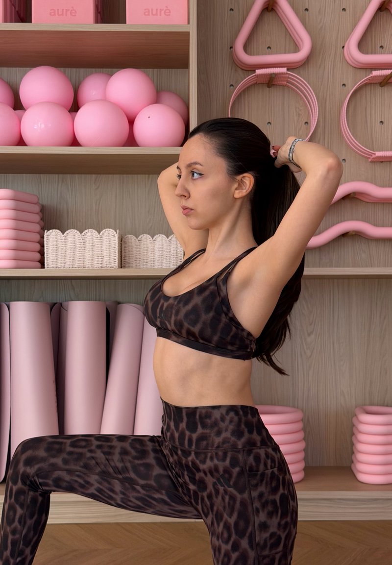 Γυναίκα με αθλητικά ρούχα λεοπάρ τυπώματος δένει τα μαλλιά της πίσω σε στούντιο γυμναστικής με ροζ γυμναστικές μπάλες, στρώματα και εξοπλισμό σε ξύλινα ράφια.