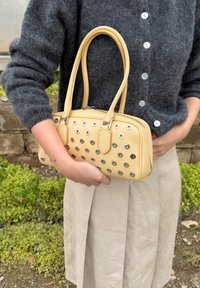 Personne portant un pull gris boutonné et une jupe beige tient un sac à main jaune avec des détails en œillets argentés en extérieur.