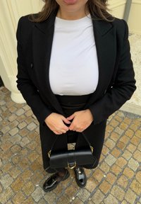 Blazer nero su una camicia bianca, abbinato a una gonna nera. Gli accessori includono una piccola borsa nera con hardware dorato e mocassini neri in vernice.