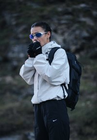 Persoon met een zonnebril, handschoenen en een licht jack die buiten de handen opwarmt met een rugzak tegen een vage natuurlijke achtergrond.