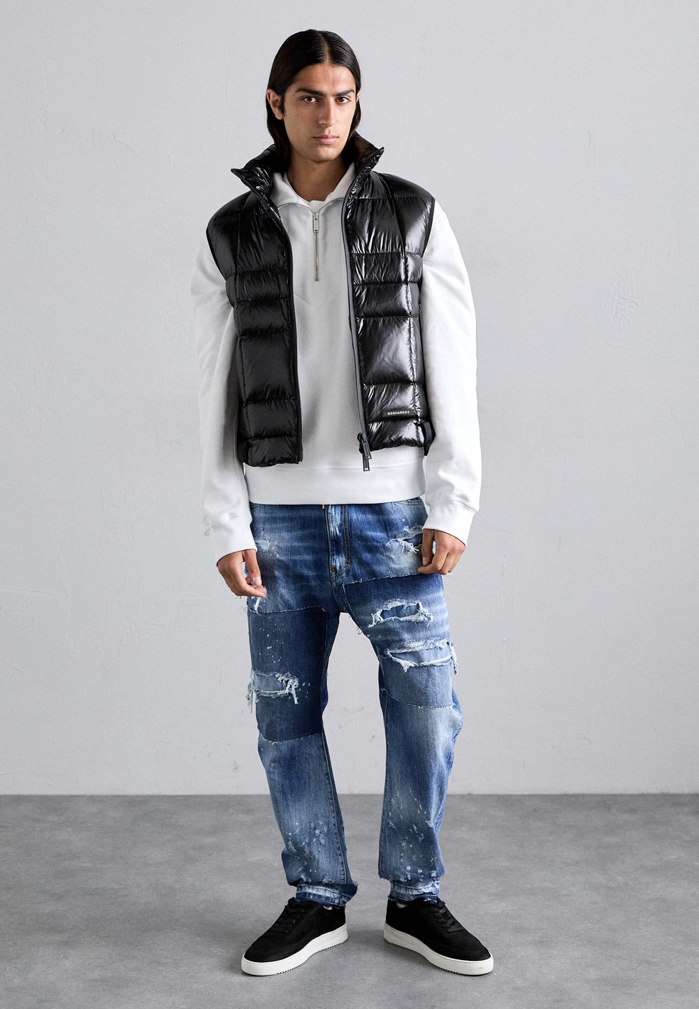 Svart vadderad väst över en vit hoodie, kombinerad med slitna blå jeans och svarta sneakers. Lätta och glänsande material, modern passform.