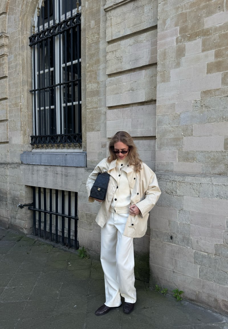 Γυναίκα που φοράει μπεζ σακάκι, λευκό πουλόβερ, λευκό παντελόνι και σκούρα παπούτσια, στέκεται κοντά σε πέτρινο τοίχο κρατώντας μαύρη τσάντα, κοιτάζει προς τα κάτω και χαμογελάει.