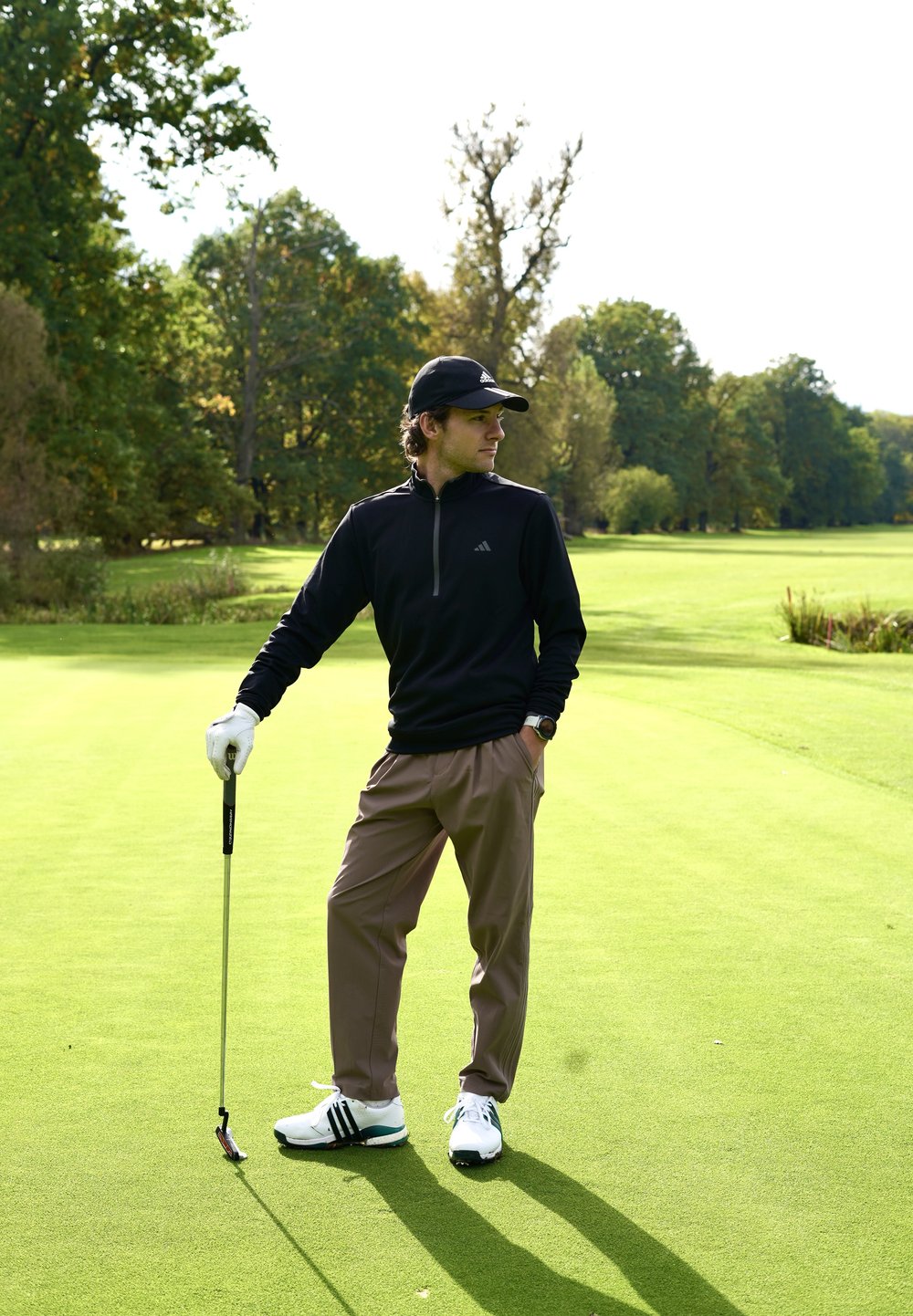 Črni golf pulover z zadrgo, bež golf hlače, beli čevlji s črnimi poudarki, drži golf palico na zelenem igrišču z drevesi v ozadju.