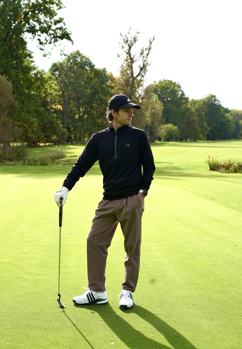 Čierny golfový pulóver na zips, béžové golfové nohavice, biele topánky s čiernymi prvkami, držiaci golfovú palicu na zelenej ferveji s stromami v pozadí.