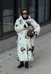 Fehér bolyhos kabát barna foltokkal, fekete ruházat és kesztyű fölött viselve. A megjelenést fekete csizmák és egy kis barna kézitáska egészíti ki.