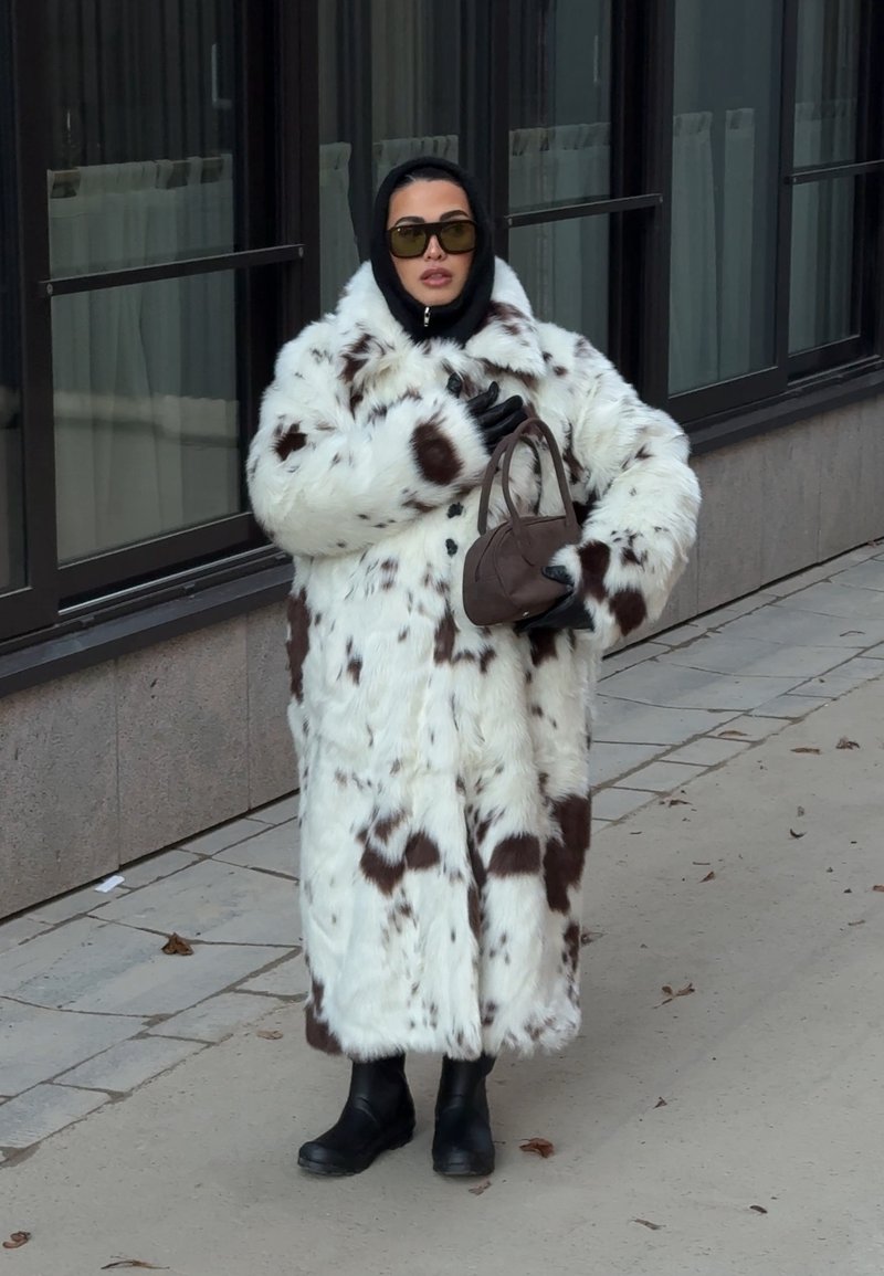 Witte zachte jas met bruine vlekken, gedragen over een zwart ensemble en handschoenen. Zwarte laarzen en een kleine bruine handtas maken de look af.