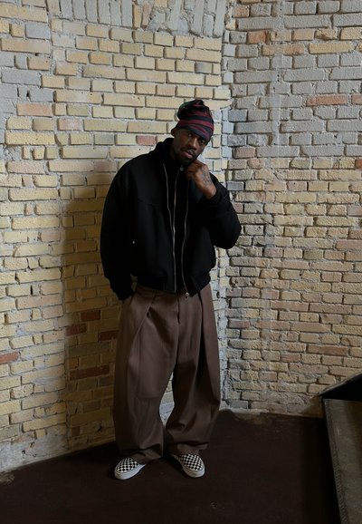 Hombre con pañuelo de rayas en la cabeza, chaqueta negra, pantalones anchos marrones y zapatos sin cordones a cuadros, de pie contra una pared de ladrillos beige.