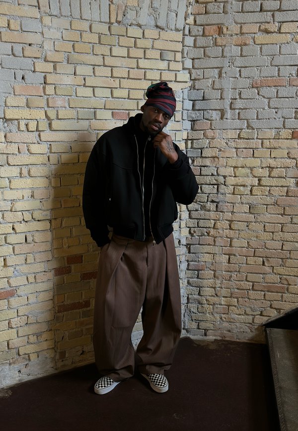 Muž s pruhovaným šátkem na hlavě, v černé bundě, širokých hnědých kalhotách a kostkovaných pohodlných botách stojí opřený o béžovou cihlovou zeď.