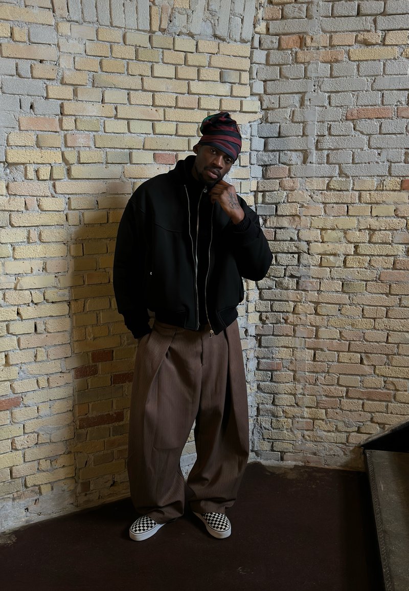 Homme portant un foulard à rayures, une veste noire, un pantalon large marron et des chaussures à enfiler à carreaux, debout contre un mur en briques beige.