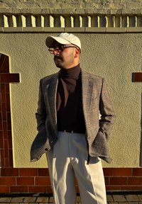 Blazer xadrez castanho sobre um suéter de gola alta escura, calças brancas, boné creme e óculos de sol. Parede texturizada com detalhes em padrão ao fundo.