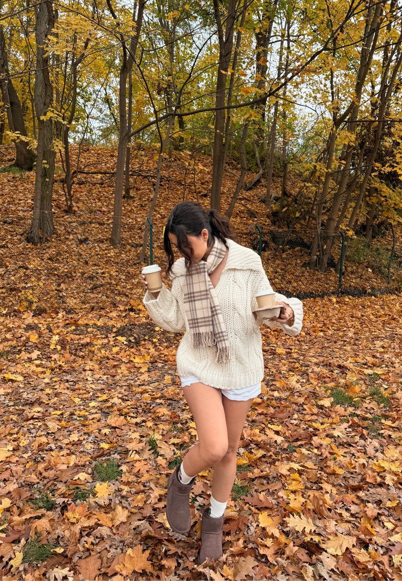 Suéter de malha creme oversize, calções brancos, botas cinza até ao tornozelo com meias claras, segurando duas chávenas de café, rodeada por folhas de outono.
