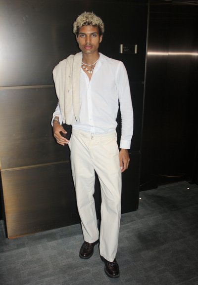 Camisa blanca de botones, pantalones crema, zapatos marrones, sosteniendo un suéter de color claro. Accesorios sencillos, cabello texturizado, frente a un fondo oscuro.