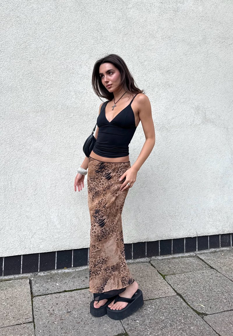 Jeune femme portant un débardeur noir, une longue jupe imprimée animalier, des sandales à plateforme noires, et portant un petit sac à main noir près d'un mur blanc.