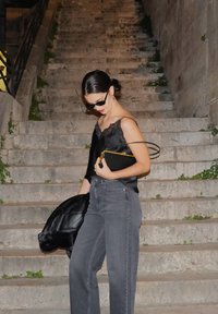 Camisola de satin preta com rendas, jeans cinzentos de cintura alta, segurando uma bolsa preta com detalhes em dourado, a pôr-se em escadas de pedra com vegetação.