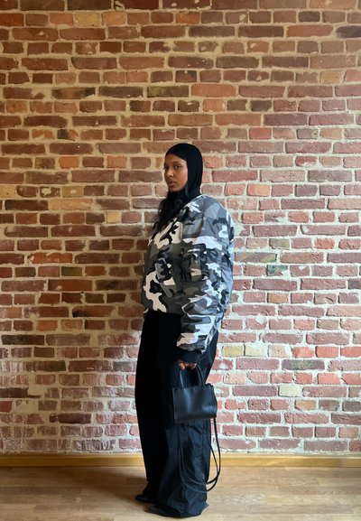 Chaqueta bomber de camuflaje en gris y blanco, pantalones anchos negros, sosteniendo un pequeño bolso negro. Pared de ladrillos texturizada de fondo.