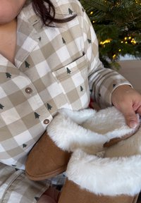 Barna papucsok fehér műszőrme béléssel, kézben tartva. Kiemelkedő kockás bézs és fehér mintájú ing zöld fa motívumokkal.