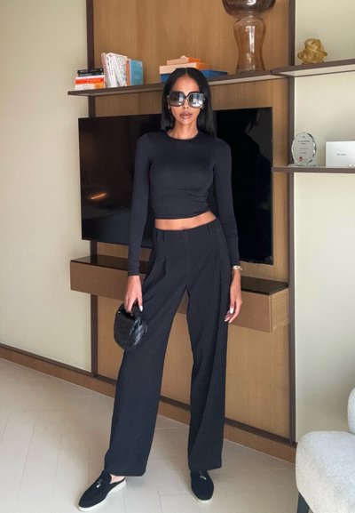 Top negro cropped de manga larga, pantalones anchos negros y zapatillas negras. La modelo sostiene un pequeño bolso negro. El fondo presenta muebles de madera.