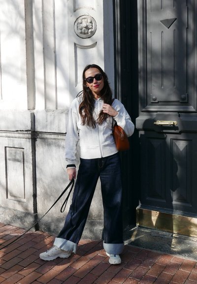 Sudadera con cremallera gris claro, jeans anchos oscuros con dobladillos, zapatillas blancas, pequeño bolso marrón, gafas de sol y fondo de pared de ladrillo.