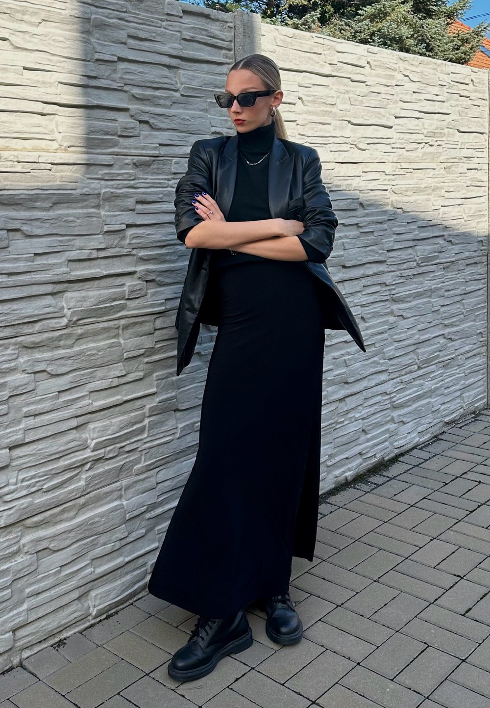 Blazer de couro preto sobre um vestido preto de gola alta, combinado com botas pretas com atacadores. O fundo apresenta uma parede de pedra texturizada.