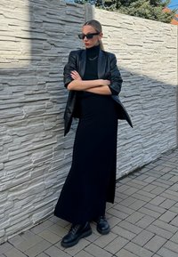 Blazer de couro preto sobre um vestido preto de gola alta, combinado com botas pretas com atacadores. O fundo apresenta uma parede de pedra texturizada.