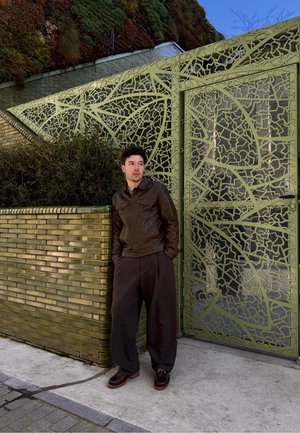 Joven con chaqueta de cuero marrón y pantalones negros apoyado en una pared de azulejos verdes junto a una puerta metálica verde con patrón.