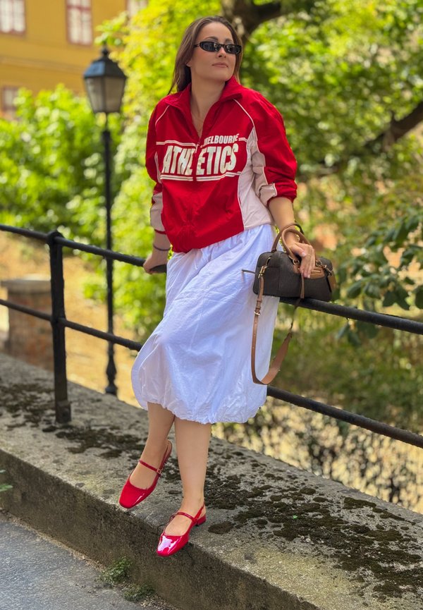 Femme portant une veste d'athlétisme rouge, une jupe blanche, des chaussures rouges et des lunettes de soleil, tenant un sac à main marron tout en s'appuyant sur une rampe à l'extérieur.