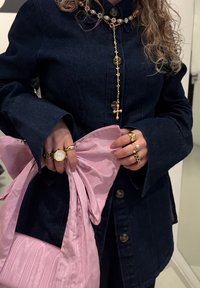 Jeansjacke mit übergroßen Ärmeln, Gold- und Edelsteinkette, verschiedenen Ringen und einer weichen rosa Satinhandtasche, die zu einer Schleife gebunden ist.