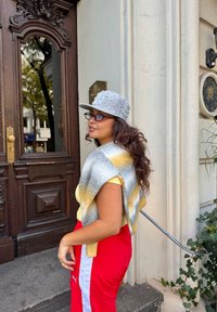 Sivi šešir s biserima, žuti i sivi prugasti šal, žuta košulja, crvene trenerke s bijelim trakama, stoji pored drvenih vrata.