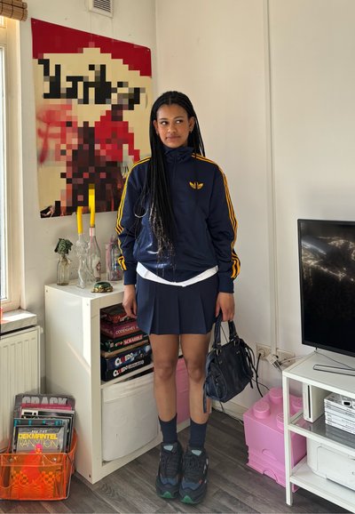 Chaqueta azul marino con cierre y rayas naranjas, camisa blanca, falda oscura con pliegues, zapatillas oscuras y un pequeño bolso negro. Entorno interior familiar.