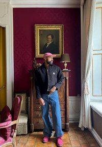 Svart kort jakke, blå jeans og rosa sko. Interiør med dype røde vegger, treverk og myke tekstiler. Merkbar lampe og kunstverk.