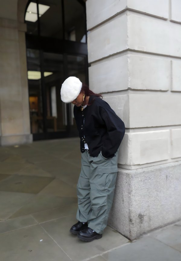 Giacca nera con bottoni, t-shirt bianca, pantaloni cargo verdi larghi, scarpe con platform nere e un cappello di pelliccia bianco, appoggiato a un muro di pietra.