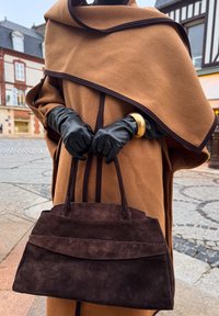 Sac à main en daim marron avec un design structuré, présentant un rabat plié et deux poignées robustes, tenu par des mains gantées sur un fond neutre.