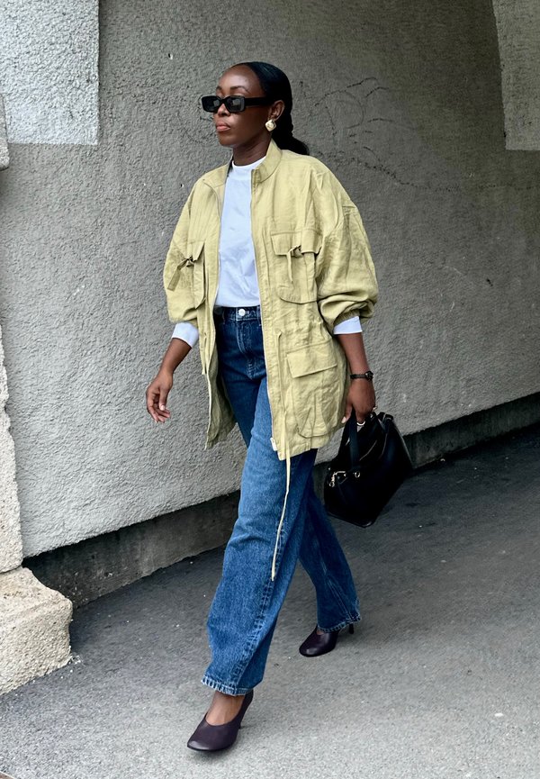 Žena nosiaca oversize béžovú bundu, biele tričko, modré rifle, čierne lodičky, slnečné okuliare a nesúca čiernu kabelku kráča okolo šedej textúrovanej steny.