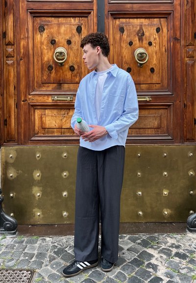 Camisa de botones azul claro, camiseta blanca debajo, pantalones anchos negros, zapatillas negras con rayas blancas, sosteniendo una botella con tapa verde.