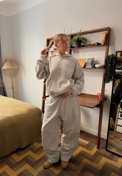 Sudadera con capucha de forro polar gris con bolsillo delantero y ajuste holgado, combinada con pantalones deportivos a juego. El modelo está de pie en una habitación con estanterías y decoración de madera.