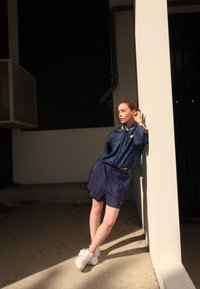 Blå og hvite stripete knapp-up skjorte og matchende shorts, parret med hvite joggesko. Modellen lener seg mot en hvit søyle i sterkt lys.