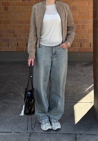 Jacke de bomber em camurça castanha clara, blusa de malha branca, jeans de perna larga cinza claro e ténis cinzentos. Saco preto segurado com uma das mãos.