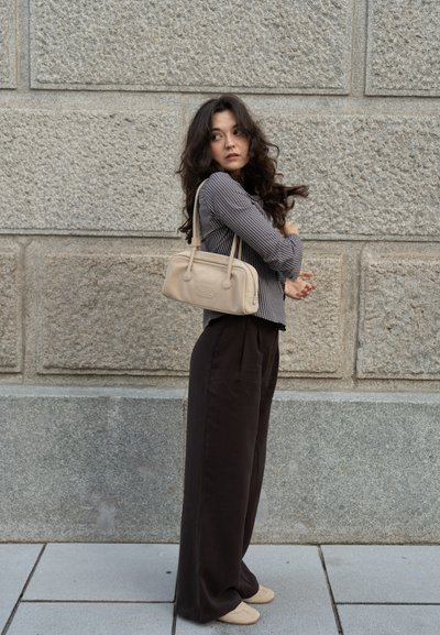 Bolso beige con dos asas, de textura en tela, combinado con una camiseta de rayas de manga larga y pantalones negros holgados, frente a una pared de piedra.