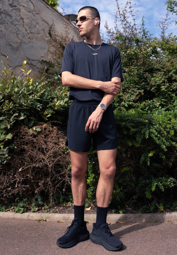 Jonge man met een zwarte sportoutfit, zonnebril en oordopjes staat buiten bij struiken en een stenen muur en kijkt opzij.