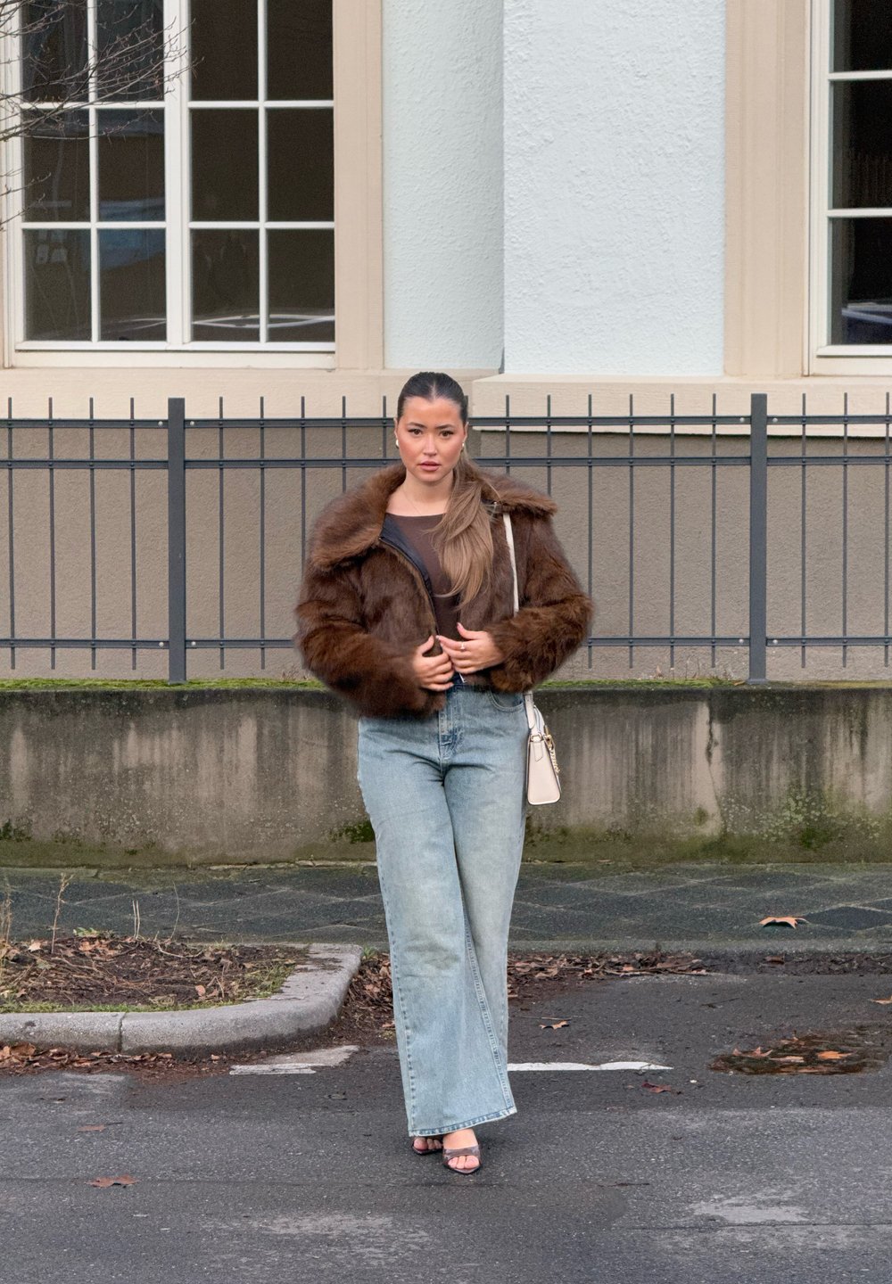 Giacca in pelliccia sintetica marrone con cappuccio, abbinata a jeans a gamba larga blu chiaro. È visibile anche una piccola borsa bianca e scarpe aperte nere.