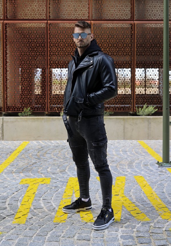 Črna usnjena motoristična jakna preko črne kapuce, v kombinaciji z raztrganimi črnimi jeans hlačami in črnimi supergami z belimi podplati, stoji na cobblestone.