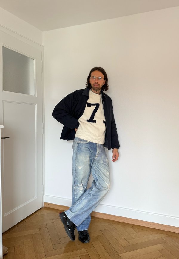 Hombre con gafas, vestido con chaqueta negra, suéter blanco con el número 7, vaqueros azules desgastados y zapatos negros, apoyado contra una pared blanca en el interior.