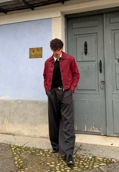 Joven con chaqueta roja, suéter negro y pantalones anchos oscuros, de pie con las manos en los bolsillos junto a una pared azul y beige y una puerta gris.