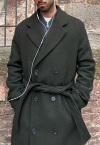 Homme portant un manteau vert foncé croisé avec ceinture, les mains dans les poches, et des écouteurs blancs, devant un mur en pierre texturé.