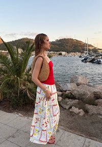Czerwona dopasowana bluzka połączona z białą, wzorzystą maxi spódnicą z owocowymi i kwiatowymi motywami; pleciona jasnobrązowa torba i czerwone sandały; nadmorskie tło.