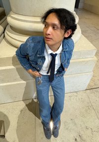 Jeans jakna čez belo srajco in modro kravato, v kombinaciji z širokima modrima kavbojkami in sivo obutvijo. Ključna veriga vidna v roki, stoji blizu stebra.