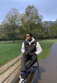 Black padded gilet over a white hoodie, black trousers, and black trainers with white accents. Sitting on a park bench in a green space.